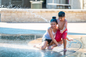 Two children in summer clothes laugh and play together in a splash pad, kneeling and standing as water jets spray around them on a sunny day.