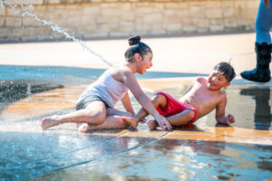 Two children play and laugh together on a wet surface as water sprays over them on a sunny day. One child sits while the other reclines, both enjoying the splash pad outdoors.