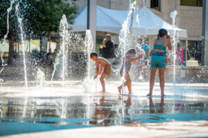 Three children play in a splash pad with streams of water shooting up around them on a sunny day. A few adults and a large tent are visible in the background.