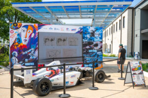 A white race car with colorful graphics is displayed outside, next to a booth promoting AI and robotics, with a person walking nearby and trees and buildings in the background.