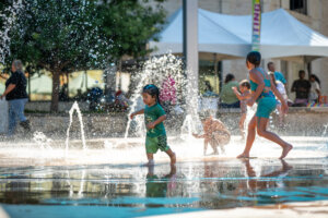 Children play and splash in a public water fountain on a sunny day. Water sprays up around them as they run and enjoy the outdoor fun. Adults and tents are visible in the background.
