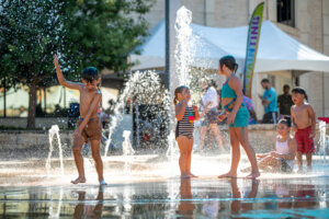 Children play and splash in a fountain on a sunny day, surrounded by water jets, tents, and people in the background.