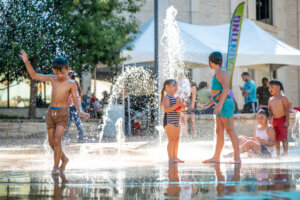 Children in swimsuits play and splash in a water fountain on a sunny day, with some laughing and others running through the spray. Adults and tents are visible in the background.