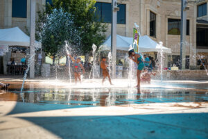 Children play and run through water fountains in a sunny outdoor plaza, with tents and a tree in the background. The water splashes create a lively atmosphere on a bright day.