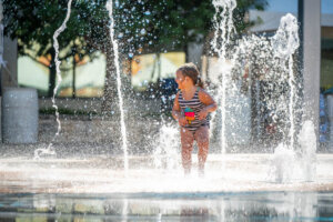 A young child in a striped swimsuit plays joyfully in a fountain, surrounded by streams of water on a sunny day. Trees and buildings are visible in the blurry background.