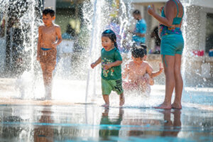 Children play and run through spraying water fountains on a sunny day, with reflections on the wet ground and splashes all around.