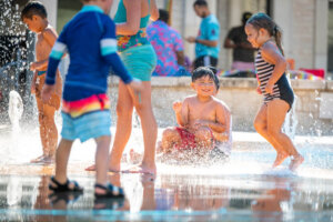 Children play in swimsuits at a splash pad, with water spraying around them. One child sits and smiles in the center, while others walk and play nearby under the sun.