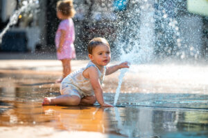 A toddler in light clothes touches water spraying from a fountain while kneeling on wet ground; another young child stands blurred in the background.