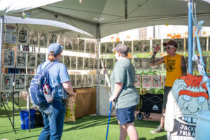 Three people look at black and white art prints displayed in a tent at an outdoor market. One person wears a yellow shirt and is gesturing. The scene is sunny with green artificial grass underfoot.