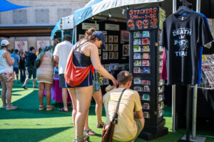 People browse sticker racks and art displays at an outdoor market with booths and merchandise, including a black DEATH TEAS T-shirt hanging on display. Bright, sunny day with green artificial turf.