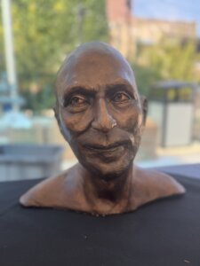 A realistic bust sculpture of a bald person with detailed facial features, set on a black surface with an outdoor, blurry background of greenery and buildings.