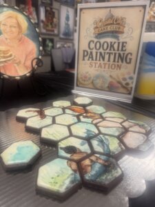 Hexagonal cookies painted with various artistic scenes are displayed on a table. A sign reads Capital City Cake Club Cookie Painting Station. A painted portrait of a woman holding a cake is in the background.