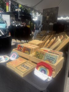 A market stall displays wooden boxes filled with colorful coasters, small rainbow crochet items, and $25 price tags under a white tent. T-shirts hang in the background.