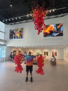 A smiling person in a tie-dye shirt holds red and yellow balloon sculptures in an art gallery, under a matching hanging sculpture, with people and artworks visible in the background.