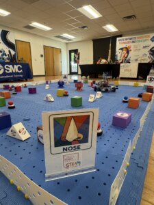 A blue pegboard arena with colorful blocks and robots set up for a STEAM robotics competition. A sign labeled NOSE with a graphic is in the foreground. Banners and tables are in the background.