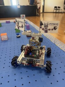 A small wheeled robot with circuit boards and flags reading PITSCO sits on a blue perforated mat in a room with tables, chairs, and colorful blocks.