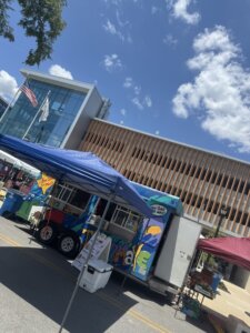 A colorful food truck sits under a blue canopy at an outdoor event near a modern building with flags and a partly cloudy sky overhead.