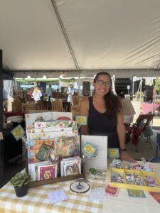 A woman stands and smiles behind a display of colorful art prints, cards, and stickers at an outdoor market booth under a white tent. The table is covered with a yellow checkered cloth and small plants.