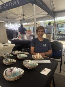 A woman sits at a booth with handmade abalone shell dishes and crystals on display, under a tent at an outdoor market. Other vendors and market tents are visible in the background.