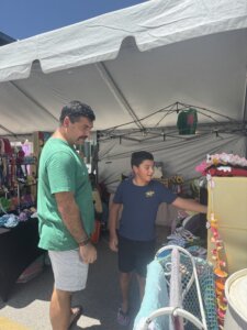 A man and a boy browse colorful handmade items at an outdoor market under a white tent on a sunny day. The boy is reaching toward a display shelf, while the man stands beside him, smiling.