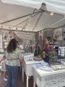 A woman wearing sunglasses speaks with a customer at a jewelry booth called Lindsay Designs at an outdoor market. Display tables hold colorful jewelry and earrings under a white canopy.