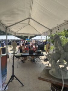 People sit under a white canopy tent at an outdoor event with art displays and sculptures, working on laptops and talking. Other tents, posters, and greenery are visible in the background on a sunny day.