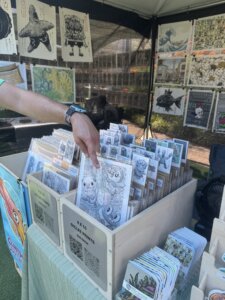 A person points to cartoon art prints displayed in wooden boxes at an outdoor art booth, with various colorful illustrations and posters hanging on the booths walls.