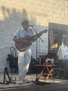 An older man in a white outfit and hat plays guitar and sings into a microphone on an outdoor wooden stage, with a microphone stand, small table, and posters behind him.