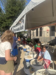Children sit at pottery wheels under a tent, shaping clay with their hands. An adult stands nearby observing. Buckets and pottery tools are on the ground, and a banner above reads Community Clay Studio..