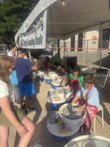 Children and adults work on pottery wheels under a white tent at an outdoor event. A few people watch and take photos, enjoying the sunny day and creative activities.