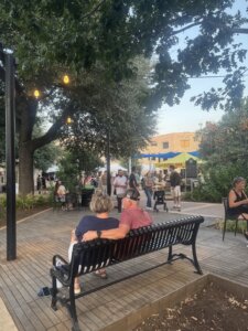 A couple sits on a bench under string lights at an outdoor market with people, food stands, and trees visible in the background.