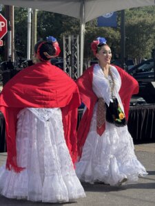 Two women in traditional white dresses with red shawls and floral headpieces dance outdoors, smiling, during a cultural celebration.