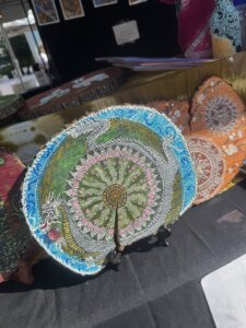 Hand-painted wooden circles with intricate mandala patterns in blue, green, and orange hues are displayed on stands at an art market booth.