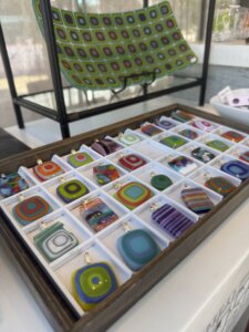 A wooden tray holds colorful square glass pendants in various patterns and hues. In the background, a large glass plate with a green and purple geometric design is displayed upright.
