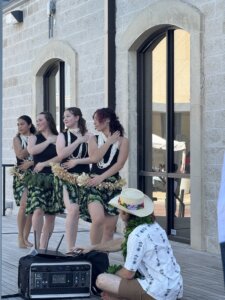 Four women in green hula skirts and white leis perform a dance on a wooden stage, while a seated man in a straw hat and floral shirt plays an instrument in front of them. They are outside a stone building.