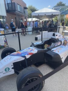 A decorated race car is displayed outdoors near a crowd and a tent. People are standing and walking around on a sunny day. The car is white with colorful art and sponsor logos.