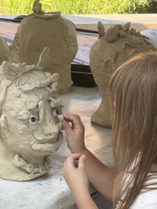 A child adds details to a clay sculpture of an animal-like face with big eyes. Several other unfinished clay sculptures are visible in the background on the table.