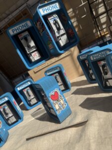 Miniature blue payphones with PHONE labels are displayed on a table, some decorated with colorful stickers, including a cartoon heart figure. Sunlight casts shadows across the scene.