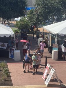 People walk through an outdoor art market with white tents displaying paintings. Some visitors examine the artwork, while others stroll in groups. A woman uses a pink umbrella for shade. Trees and signs are visible in the background.