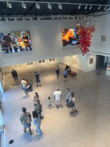 People view art in a modern gallery with large photos on the walls and a red glass sculpture hanging from the ceiling. Some visitors stand in groups while others observe artwork individually.
