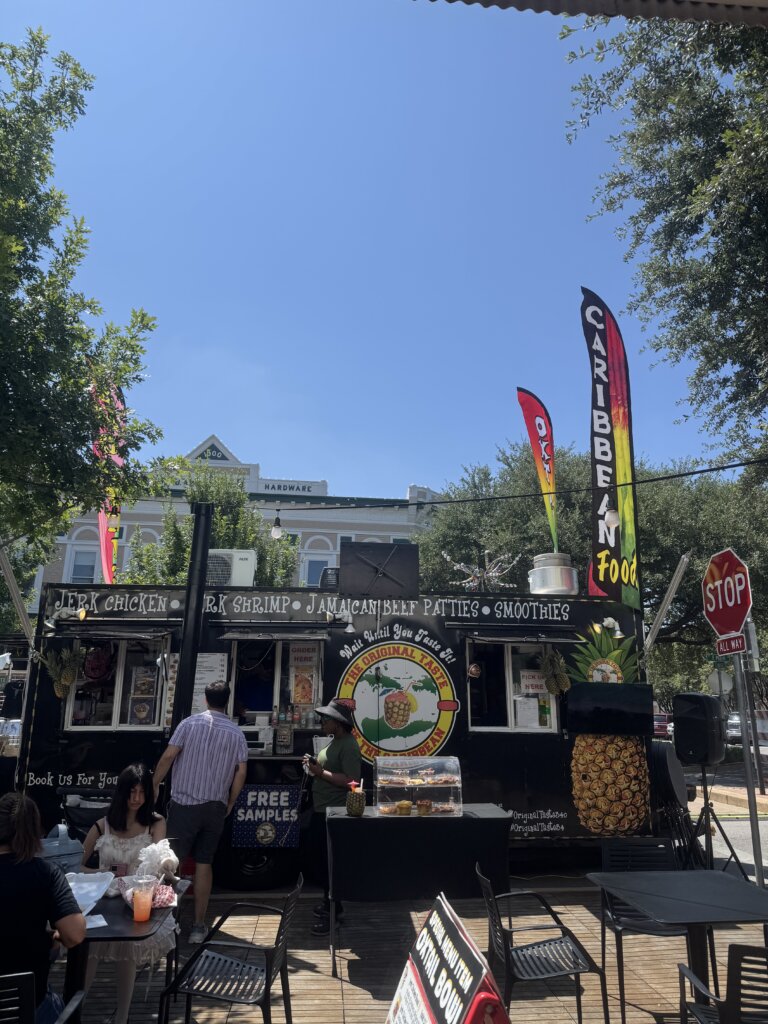 A Caribbean food truck with colorful flags is parked on a sunny street. People wait in line, and tables with chairs are set up nearby. Trees provide shade, and a stop sign is visible on the right.