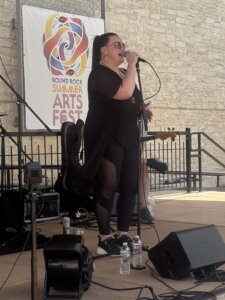 A woman sings into a microphone on an outdoor stage at the Round Rock Summer Arts Fest, wearing black clothing and sunglasses, with music equipment around her.