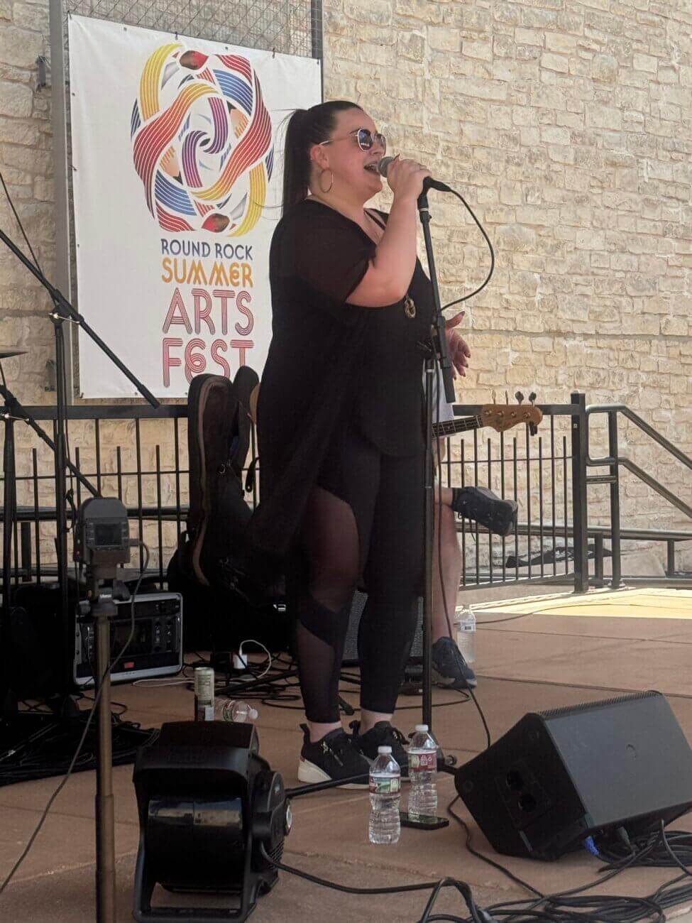 A woman in black sings into a microphone on an outdoor stage at the Round Rock Summer Arts Fest. Musical equipment and water bottles are on the ground nearby. A stone wall and festival banner are in the background.