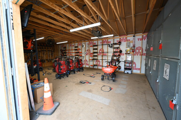 A storage shed with exposed beams, various power tools and equipment, safety cones, and shelves along the walls. The floor is mostly clear, with some items and cords scattered around.