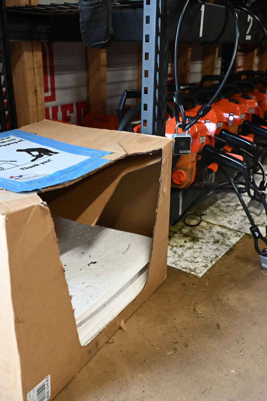 A cardboard box with a torn opening contains white absorbent pads. Behind the box, orange power tools with black cords are lined up on a metal shelf in a workshop setting.