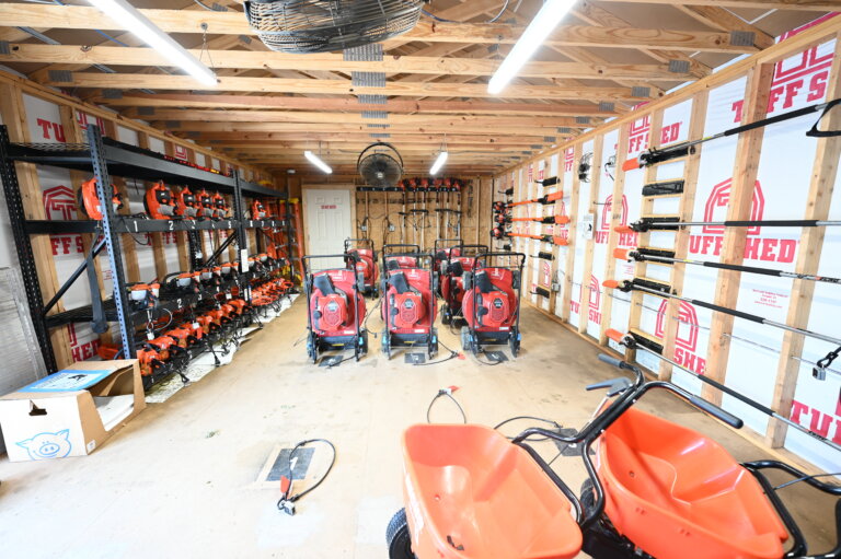 A shed interior with rows of orange lawn equipment, including mowers, trimmers, and blowers, neatly organized on shelves and the floor. Exposed wooden beams and insulation are visible on the walls.