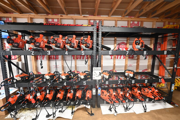 Two shelves hold multiple orange chainsaws, each numbered and organized in rows. The chainsaws are secured with cables. The background shows exposed wooden framing and construction materials.