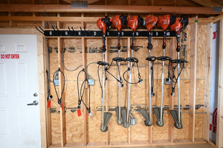 Four orange weed trimmers hang on a wall rack above five gas-powered weed trimmers hanging vertically in a wooden shed. The wall behind them is unfinished wood, and a white door is on the left side.
