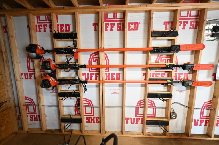 Three orange pole saws are mounted horizontally on the wooden studs of an unfinished shed wall with Tuff Shed logos printed on the white insulation panels.