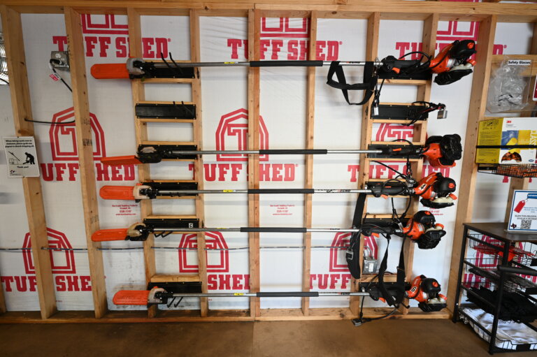 Four orange and black power trimmers are mounted horizontally on a wooden rack against a wall with Tuff Shed branding. Shelves with tools and boxes are visible on the right side of the image.